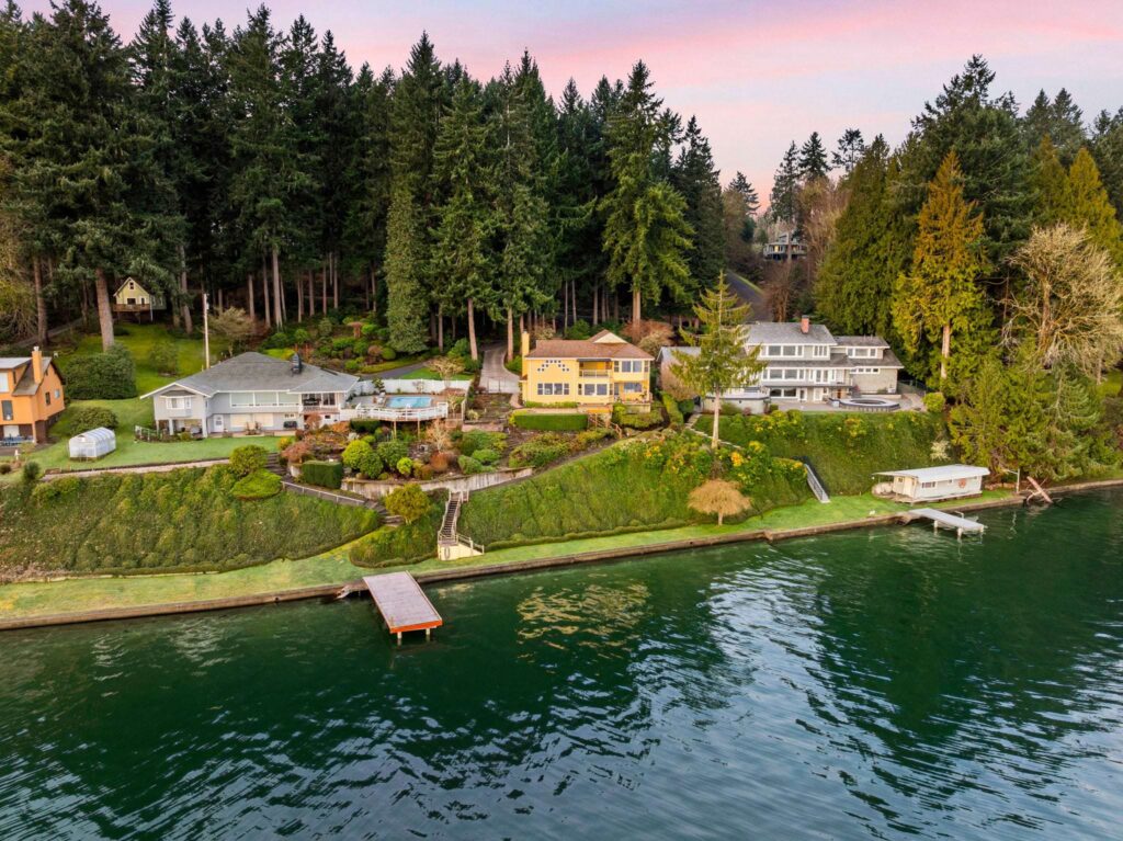 A row of houses with lush gardens sits along a waterfront, backed by tall evergreens; a dock extends into calm water beneath a sunset sky—reflecting the charm that influences Olympia WA home prices in this serene community.
