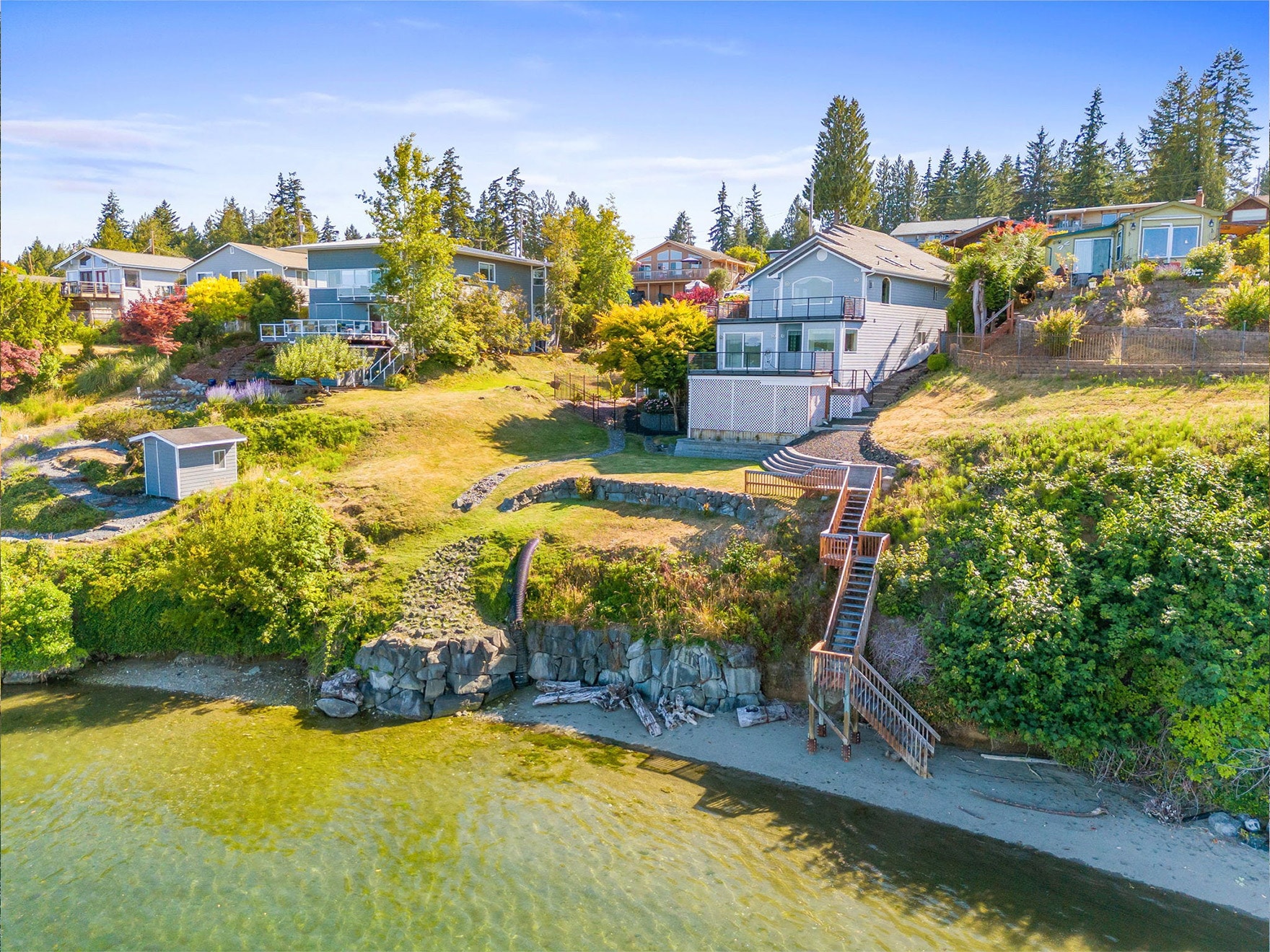 A waterfront home with large windows sits on a grassy hillside in Thurston County, where the median list price reflects its serene setting. Wooden stairs lead from the yard to a sandy shoreline along calm, clear water.