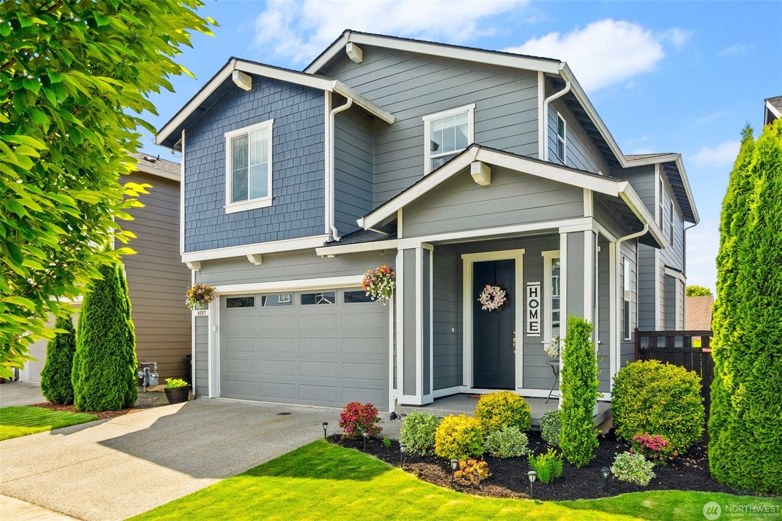 A two-story modern gray house with white trim, a double garage, well-kept landscaping, green shrubs, and hanging flower baskets, under a blue sky with scattered clouds.