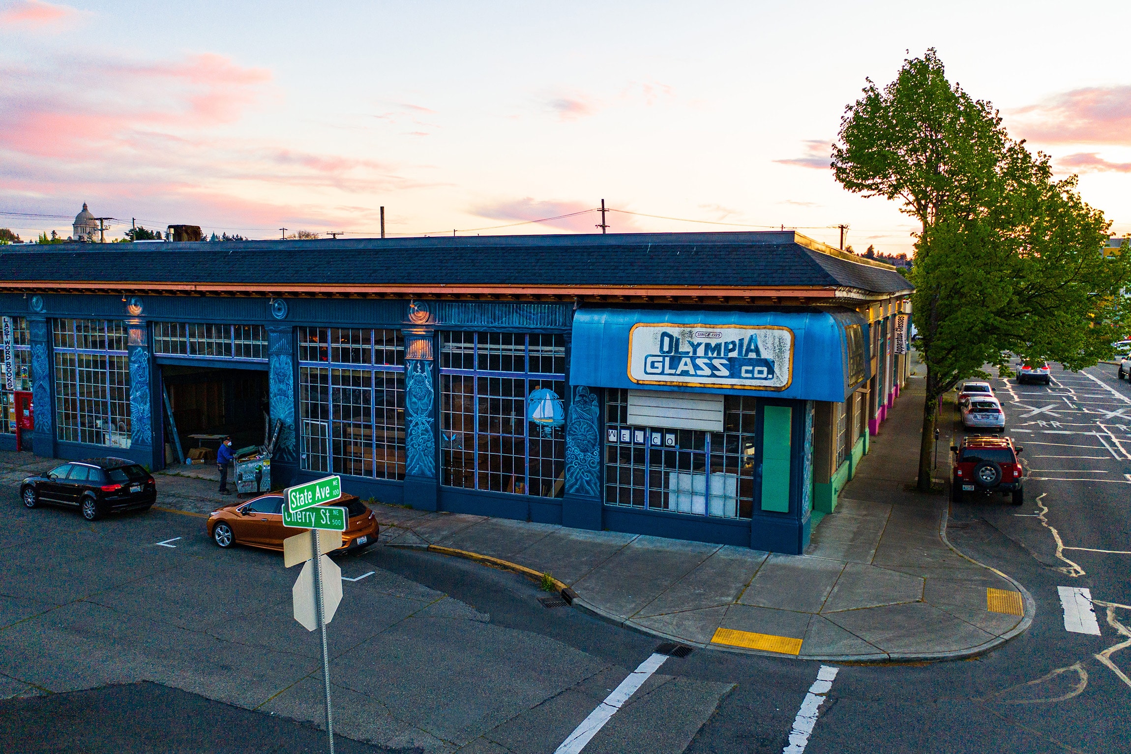A street corner view of Olympia Glass Co., a large building with many windows and a blue sign, cars parked along the street, and a tree near the sidewalk at sunset—ideal for glimpsing local life as noted in the latest Thurston County Market Report.