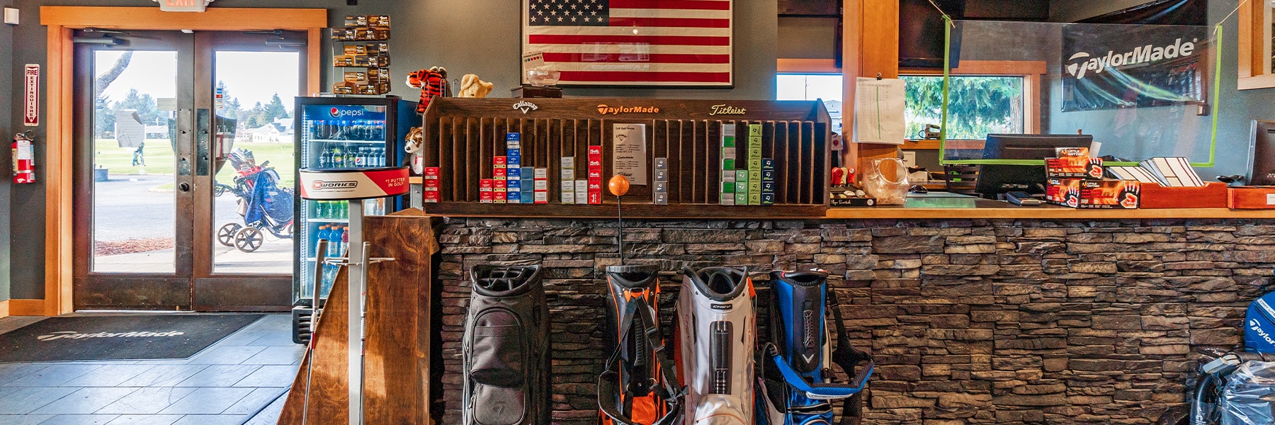 The golf shop interior boasts a stone-front counter showcasing various golf bags. Shelves brim with accessories, while an American flag adorns the wall. Beyond the door lies a stunning view of the golf course, reminiscent of the picturesque golf course homes in Thurston County.