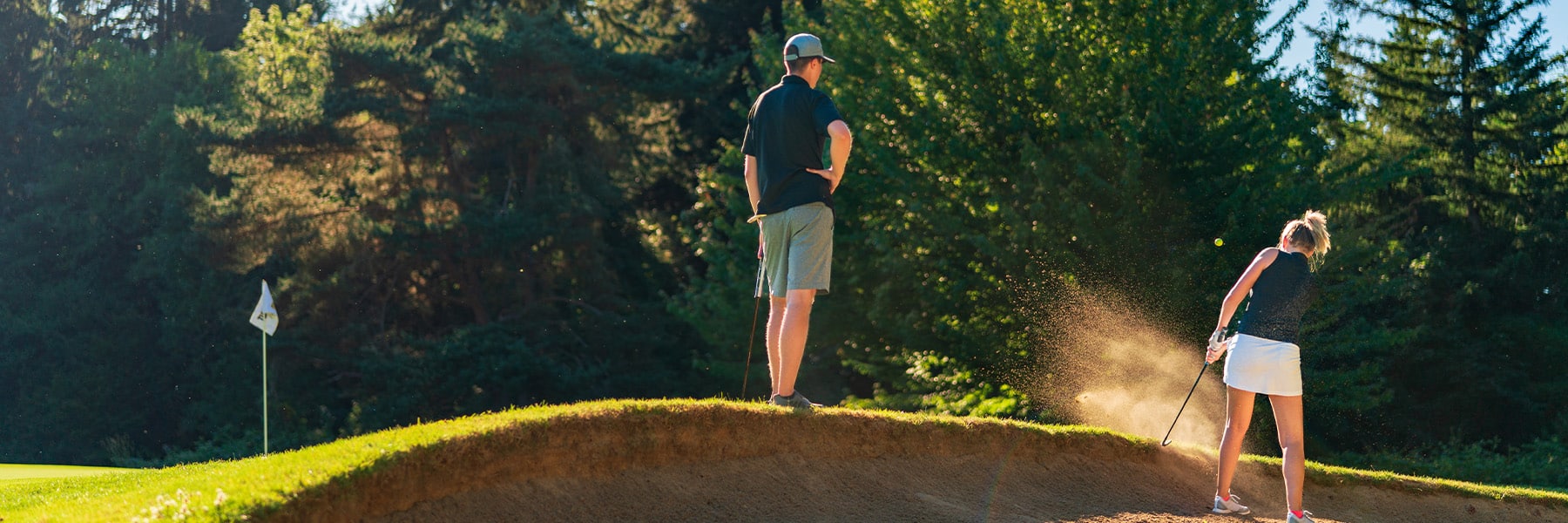 A woman swings a club in a sand trap on a sunny Thurston County golf course, sending sand flying. Nearby, a man in shorts and a cap stands, with trees and the inviting view of golf course homes and a flag fluttering in the background.