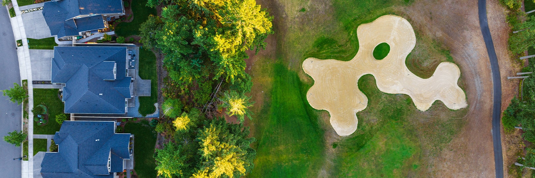 Aerial view of golf course homes in Thurston County next to a lush fairway. On the right, an irregularly shaped sand trap is surrounded by green grass. Dark-roofed houses and a road are visible on the left, with trees lining the area.