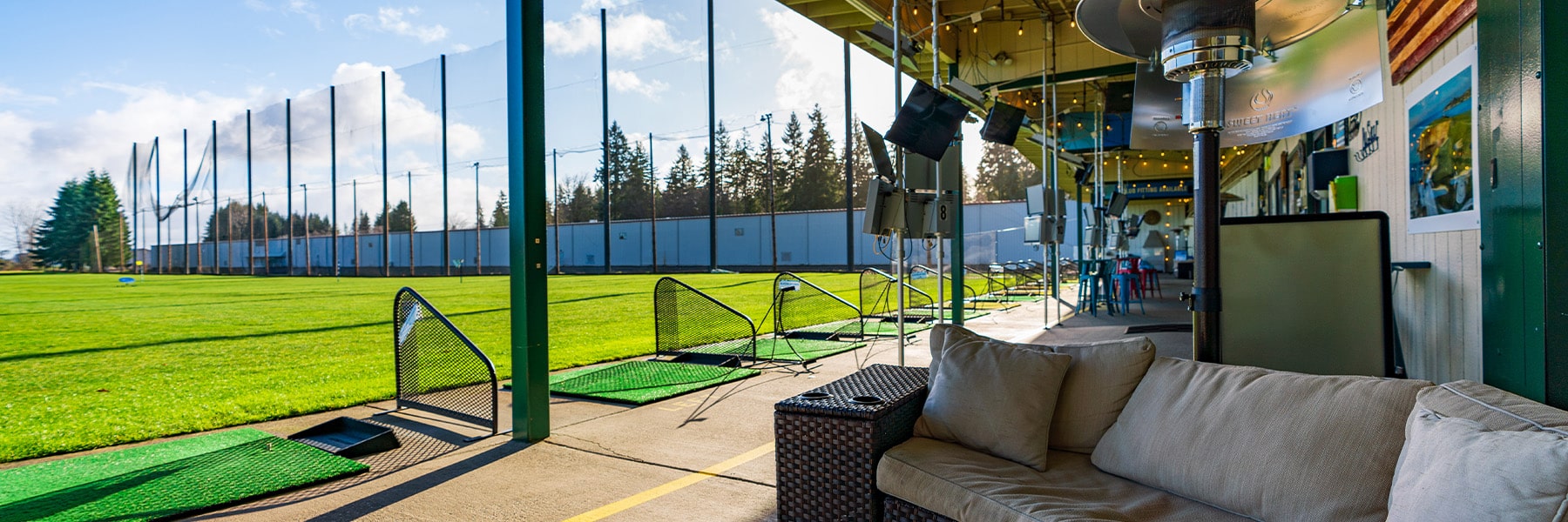 A modern golf driving range with multiple bays, each separated by mesh dividers, sits near golf course homes in Thurston County. Green mats and tees line the bays, while comfortable seating with a tan sofa graces the foreground. Trees and a clear sky provide a serene backdrop.