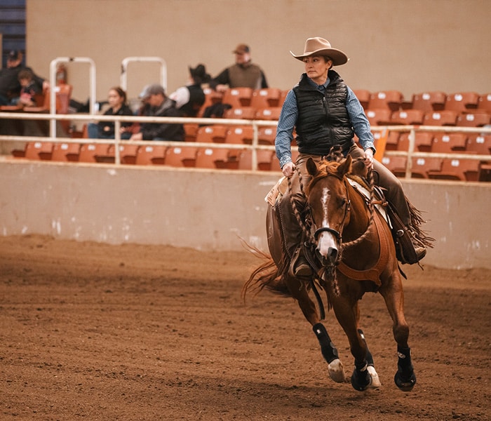 A person wearing a cowboy hat rides a brown horse in an indoor arena in Olympia, with rows of empty orange seats and a few spectators—capturing the spirit of Thurston County’s horse properties.