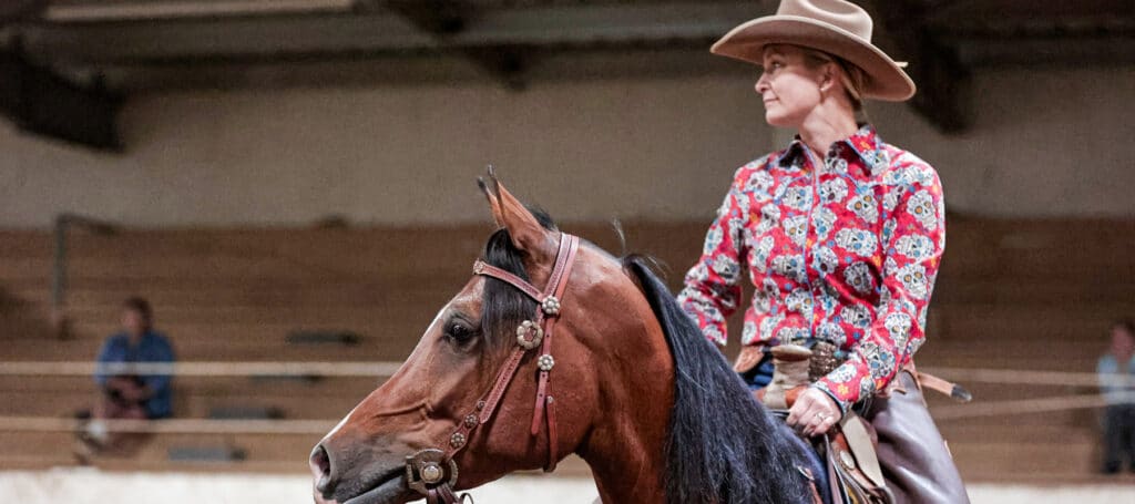 A person wearing a cowboy hat and a colorful patterned shirt rides a brown horse in an indoor arena with empty wooden bleachers, showcasing the vibrant equestrian spirit found in Olympia and Thurston County horse properties.