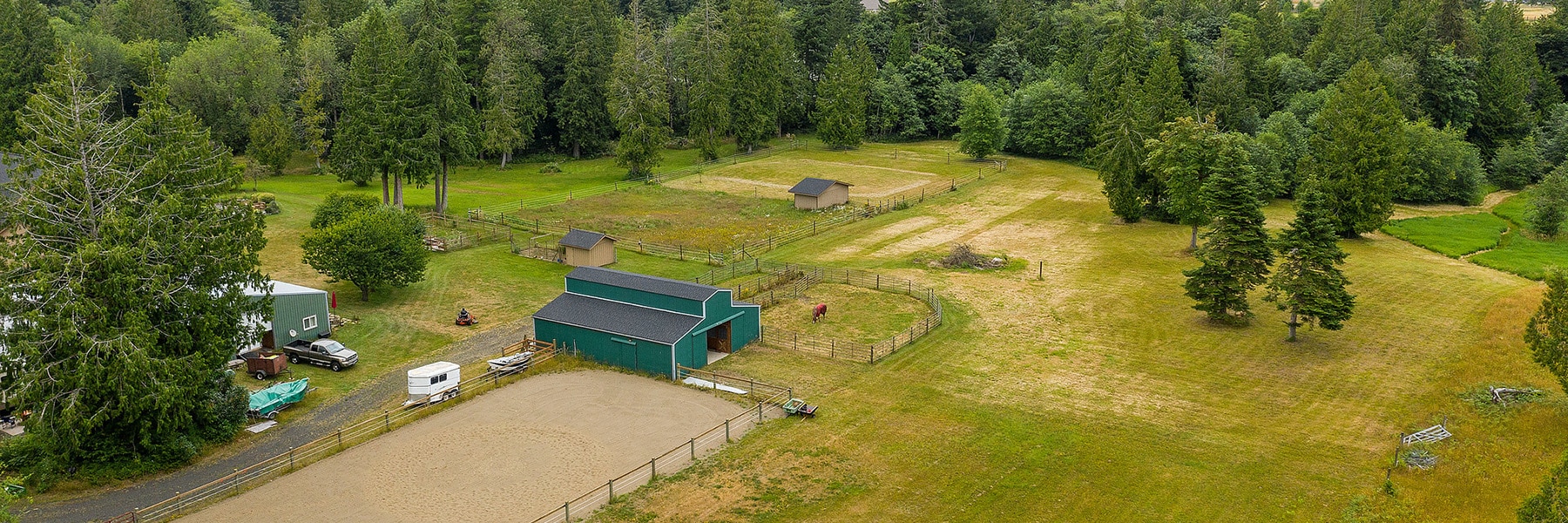 Aerial view of a rural horse property in Thurston County near Olympia, featuring a green barn, fenced riding arena, grazing horses, small outbuildings, and trees. A house, vehicles, and equipment are visible on the left side.