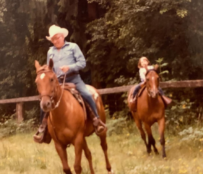 An older man in a cowboy hat rides a horse in the foreground, while a young girl follows behind on another horse. They are outdoors near a wooden fence and trees, enjoying the scenic beauty of Thurston County horse properties.
