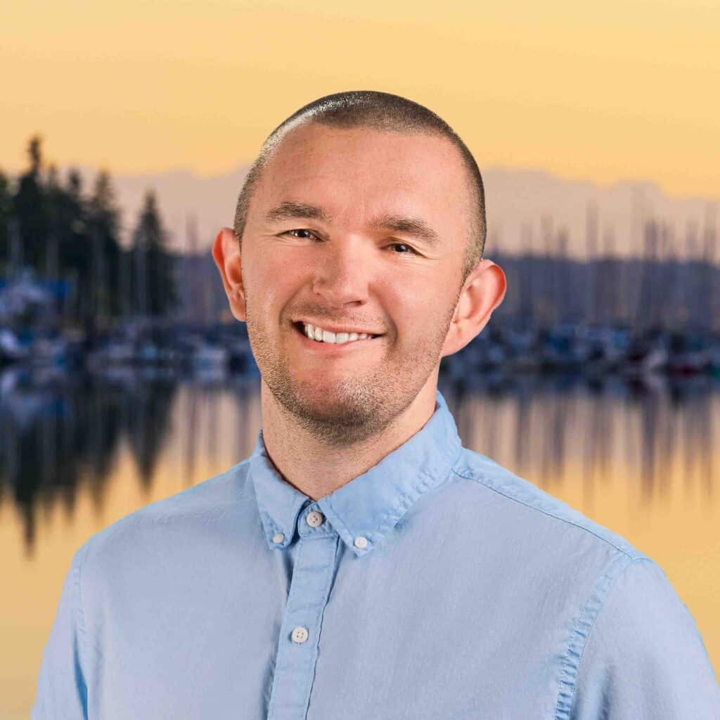 A smiling man in a light blue button-up shirt stands in front of a marina at sunset, with blurred sailboats and trees behind him, ready to discuss the latest Thurston County Market Report and current interest rates in Thurston County.