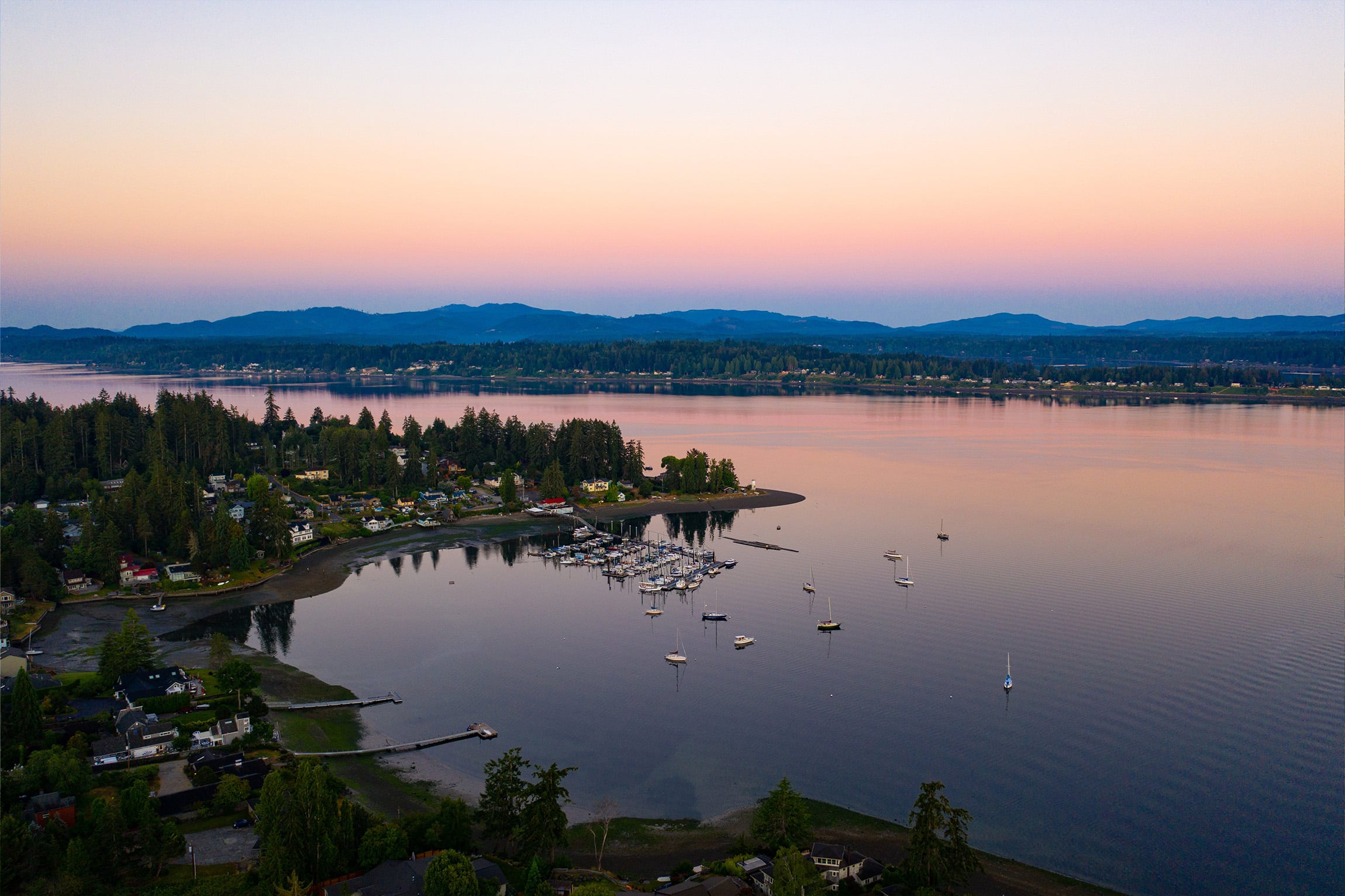 Aerial view of a calm lake at sunset with a marina, boats, and waterfront homes surrounded by trees. Soft pastel colors fill the sky, reflecting on the water, with mountains in the distance—perfect for a Thurston County saltwater report.
