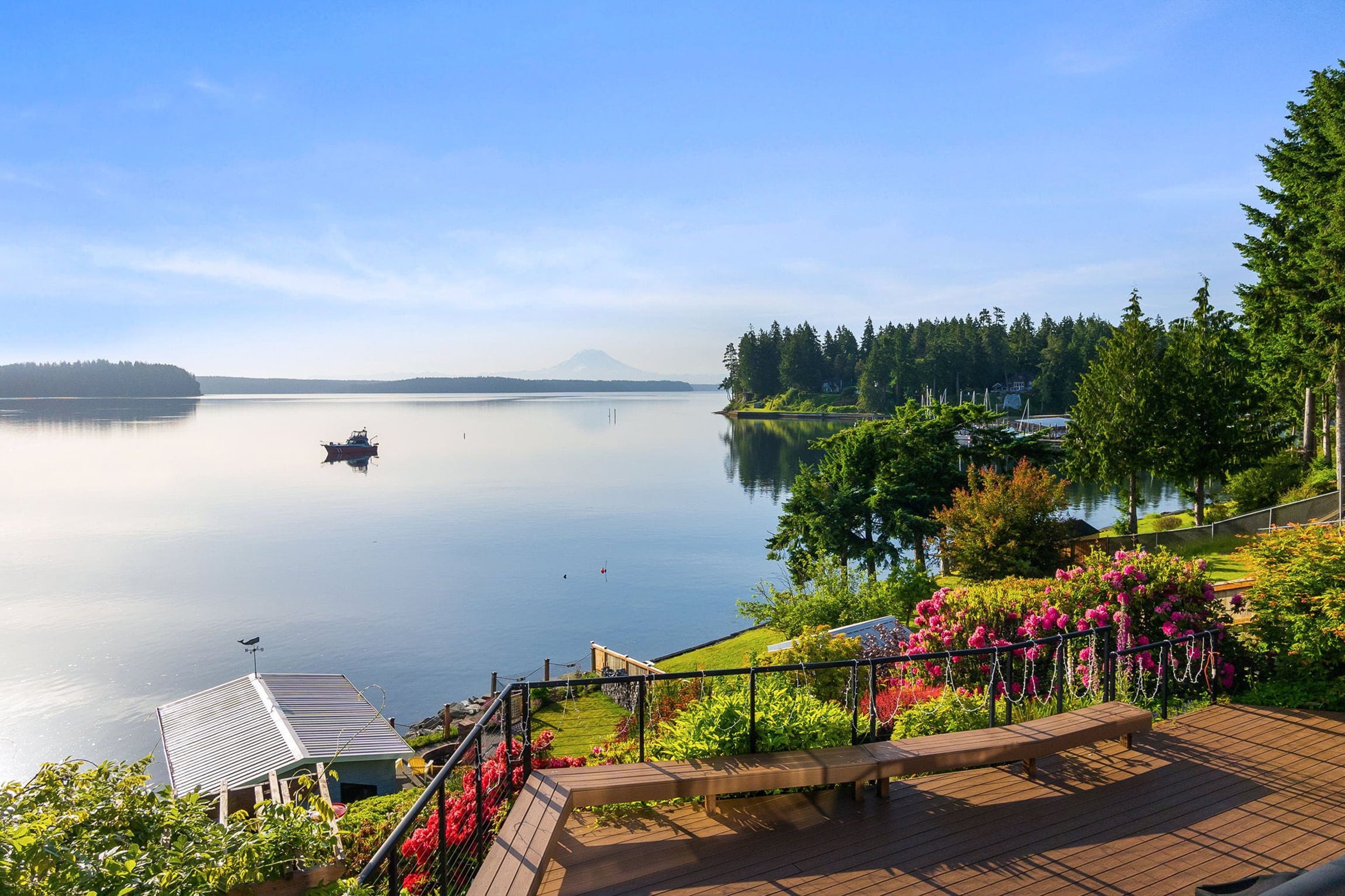 A scenic lakeside view with calm water, a dock, and a boat invites relaxation, echoing the peaceful vibes of a Thurston County saltwater report. Lush greenery, blooming flowers, and distant mountains shine under a clear blue sky.
